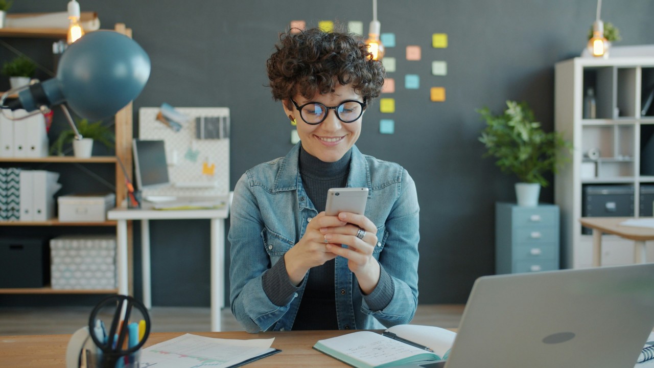 An elegant evening social gathering in a modern kitchen or dining area. Five people are mingling and conversing, dressed in smart casual attire including blazers and leather jackets. In the foreground, a wooden table displays an array of wine bottles, glasses, candles, and a potted plant, creating an intimate dinner party atmosphere. The lighting is warm and ambient, with horizontal blinds visible in the background and modern fixtures creating a sophisticated setting for the social event.